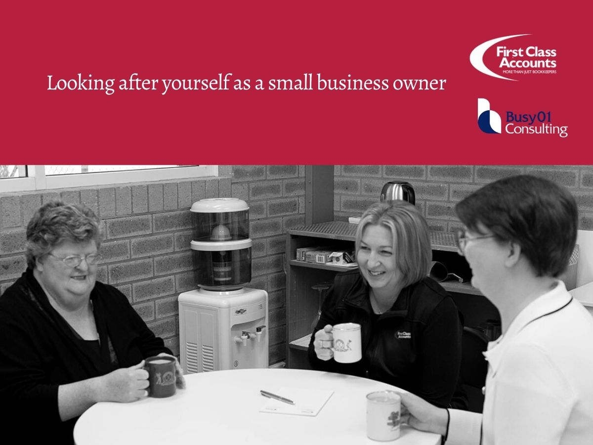 Three women sitting at a round table in an office, smiling and holding coffee mugs during a relaxed business discussion at First Class Accounts Ovens & Murray and Busy01 Consulting.