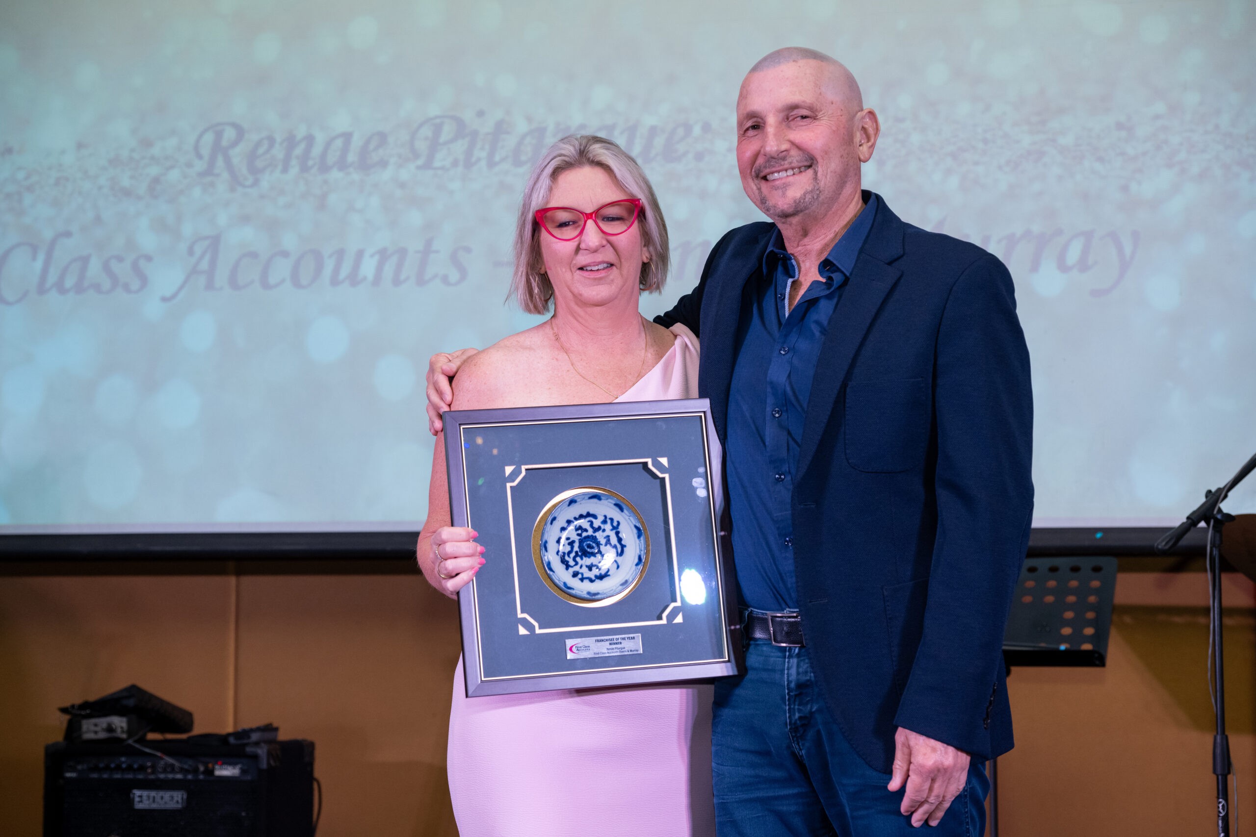 Two people standing together at an award presentation. The woman on the left is holding a framed award, and both are smiling at the camera. The background screen displays partially visible text referencing First Class Accounts Ovens & Murray.